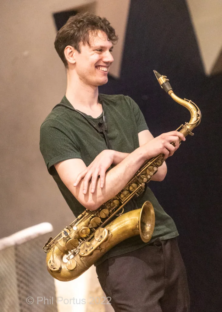 A young man with curly hair smiles while holding a brass saxophone.