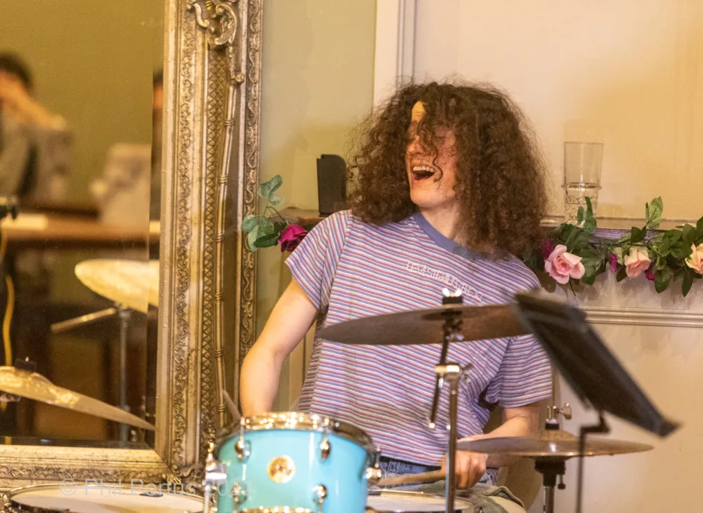 A person with curly hair plays a blue drum set while smiling and holding drumsticks.