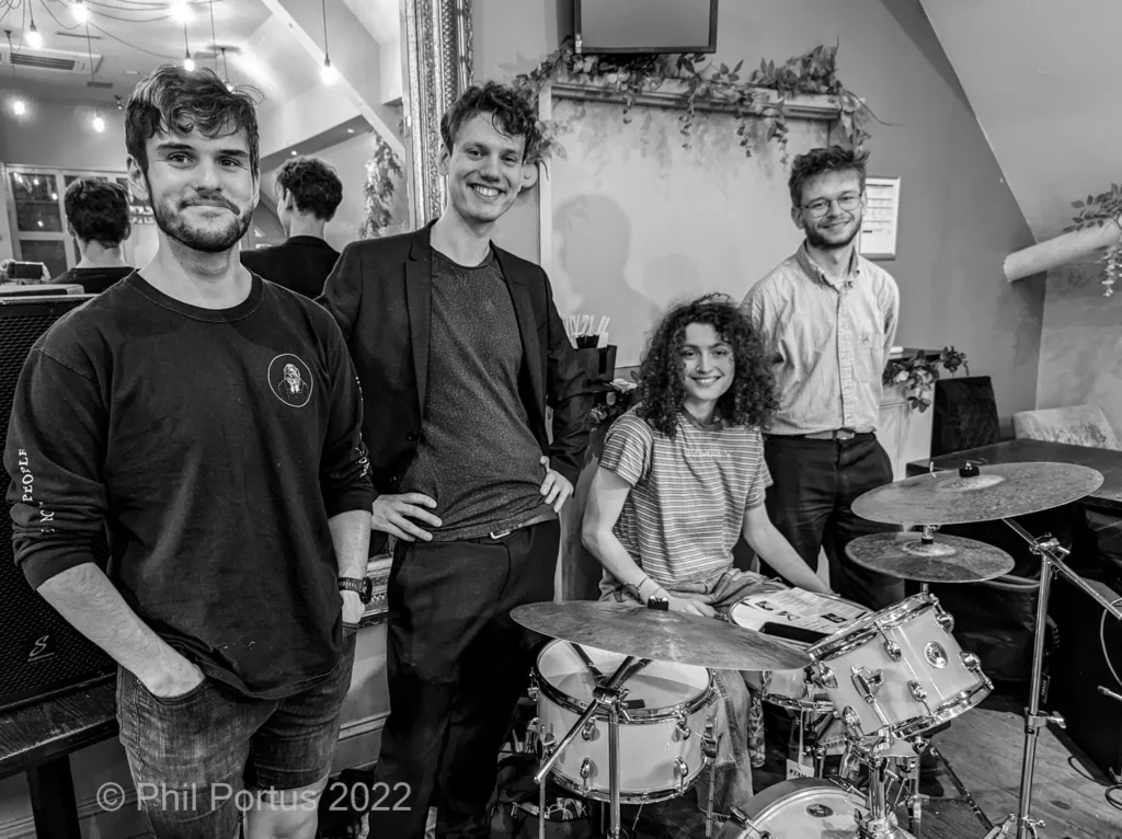 Four people stand near a drum kit in a black and white portrait.