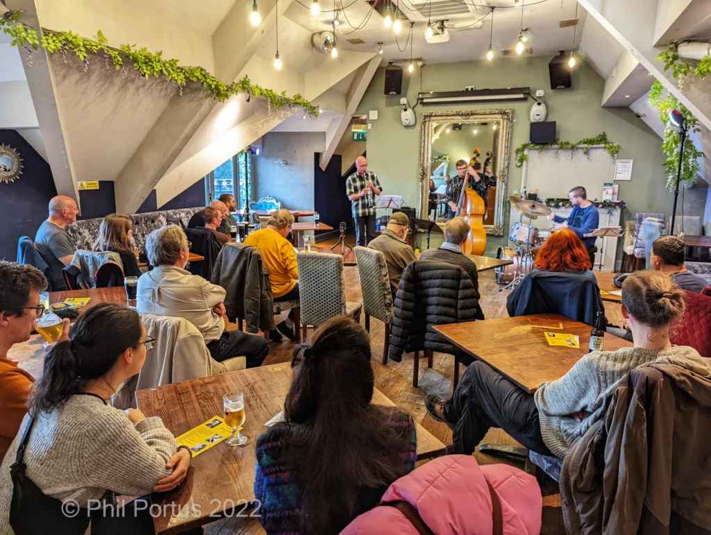 A group of people sit at tables listening to a band performing on a small stage in a dimly lit room.