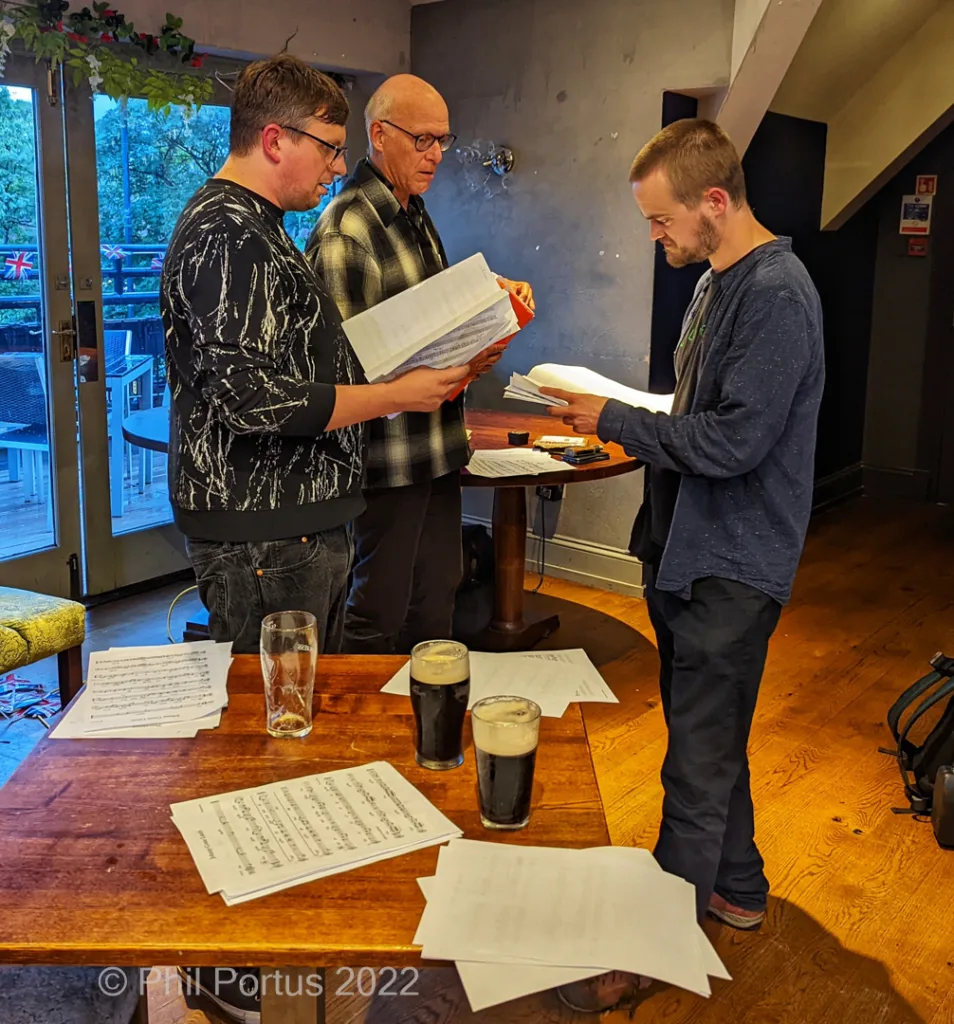 Three men examine sheet music spread across a wooden table with glasses of dark beer.