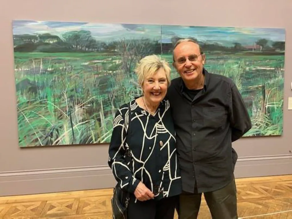Two adults stand close together in front of a large landscape painting on a neutral wall.