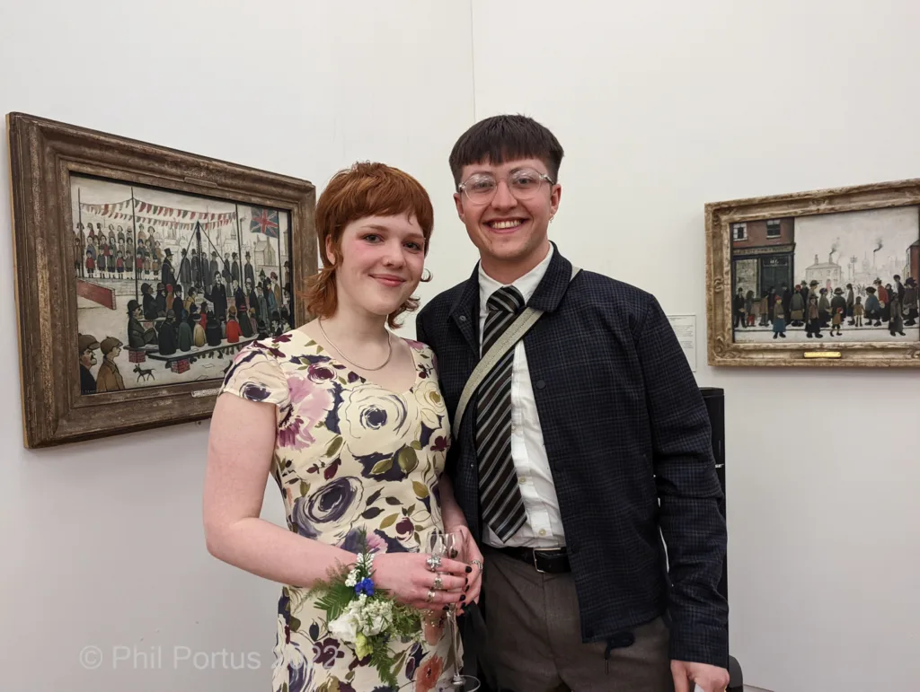 A couple poses for a picture near framed artwork in a room with white walls.