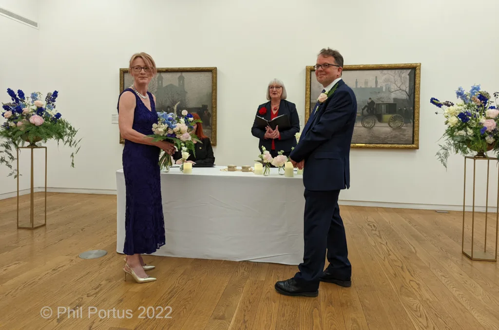 A couple stands at a table during a ceremony in a room with white walls and framed artwork.