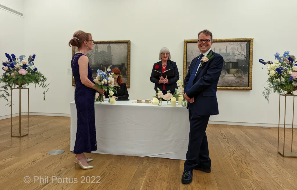 A couple stands at a table during a ceremony in a bright room with artwork on the walls.