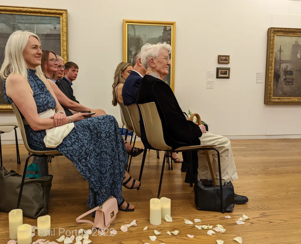 Several people sit on chairs arranged in rows within a room with framed artwork on the walls.