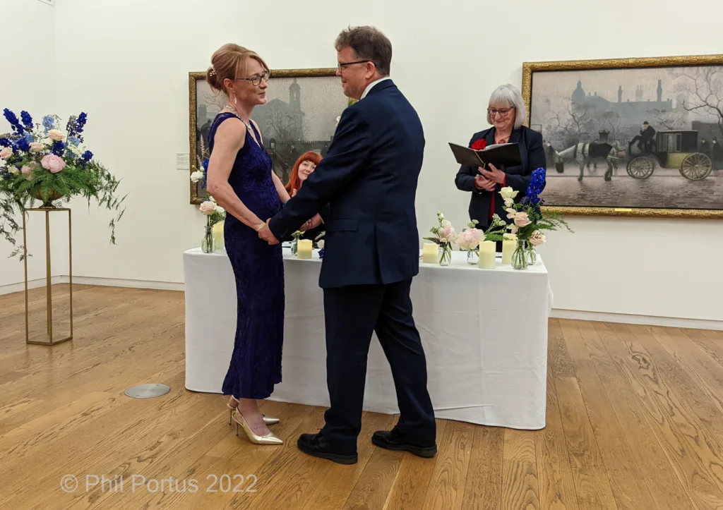 A couple holds hands at a small table during what appears to be a wedding ceremony in an art gallery.