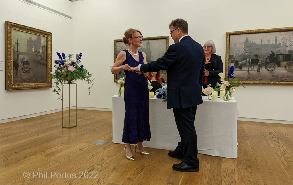 A couple exchanges vows at a table decorated with flowers in an art gallery setting.