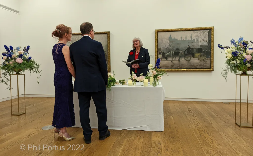 A couple stands facing a woman at a table during what appears to be a wedding ceremony in an art gallery.