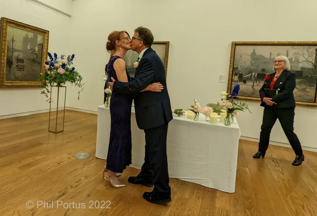 A couple embraces after a ceremony in a room with framed artwork and floral arrangements.
