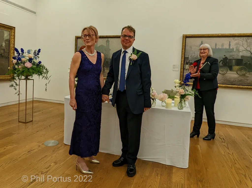 A couple stands between floral arrangements at a ceremony in a room with framed artwork on the walls.