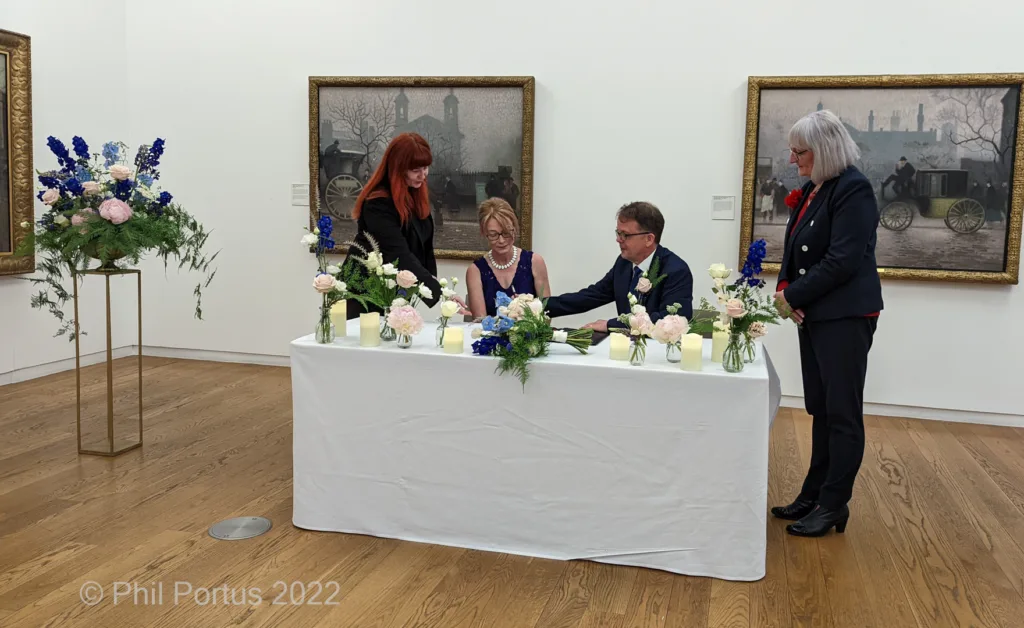 Four people stand around a white table decorated with flowers in an art gallery setting.