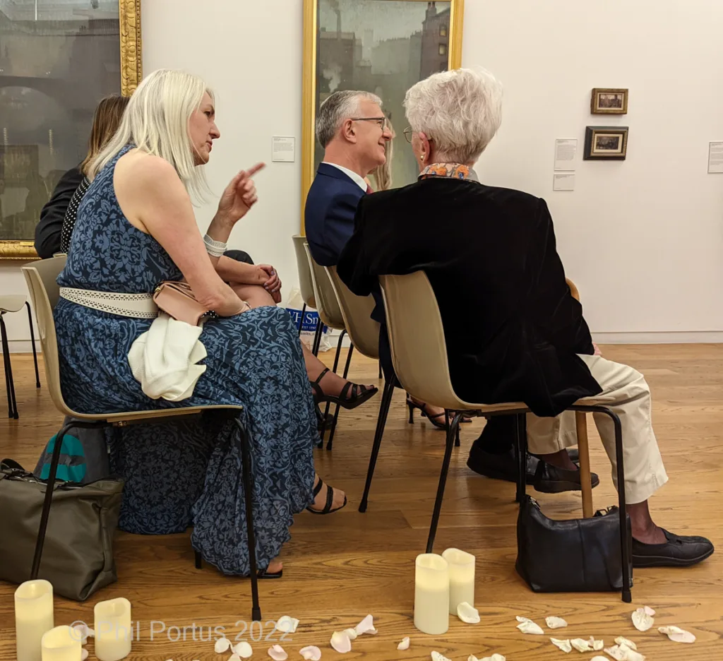 Four people sit in chairs during an event with artwork visible on the walls and candles on the floor.