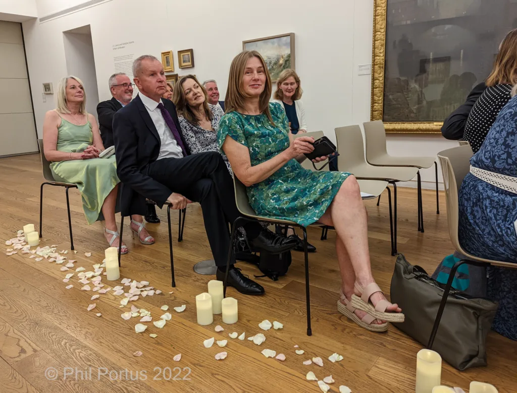 Several people sit on chairs arranged in rows within a room with artwork and candles.