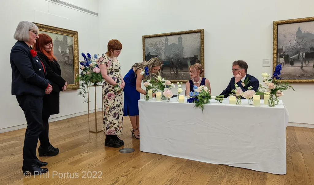 A wedding ceremony takes place indoors with several people gathered around a table decorated with flowers.