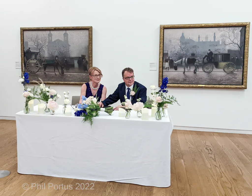 A couple sits behind a long table decorated with flowers and candles in a room with framed artwork on the walls.