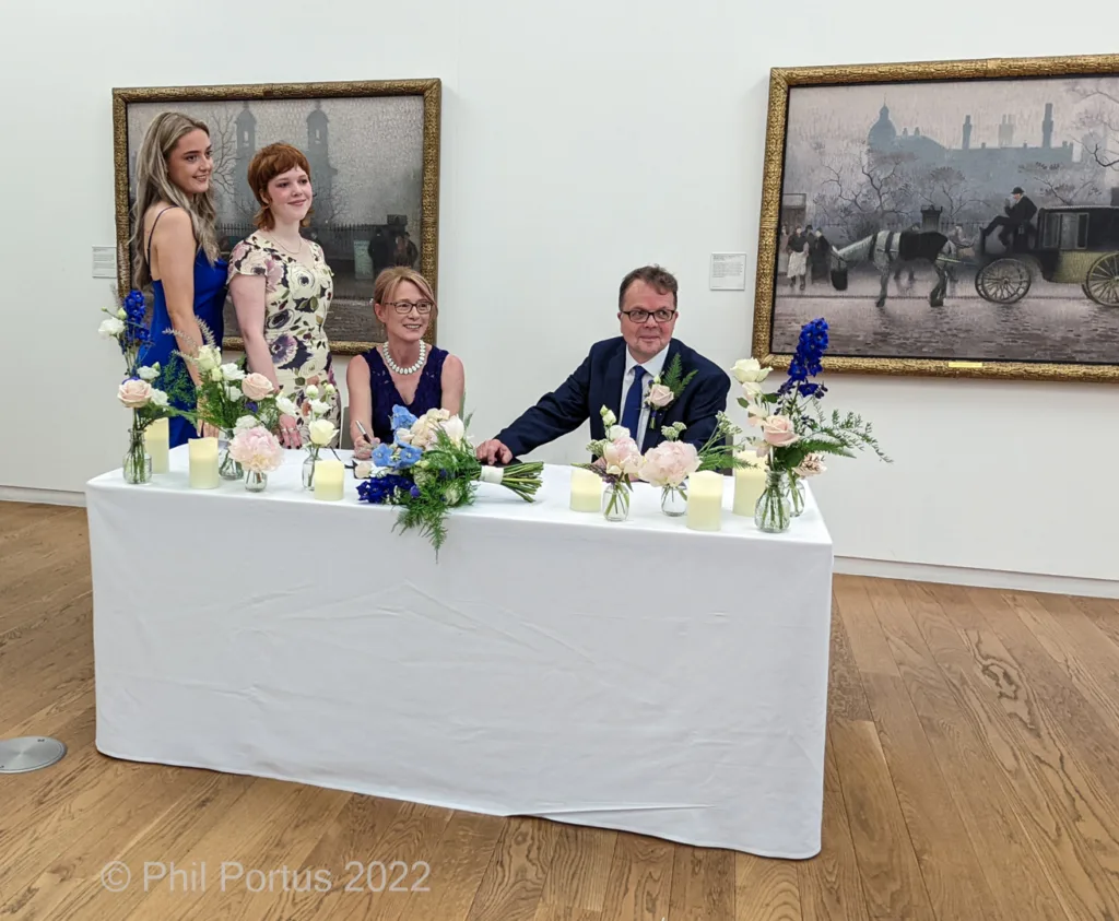 Four people stand behind a long table decorated with flowers and candles in a room with framed artwork on the walls.
