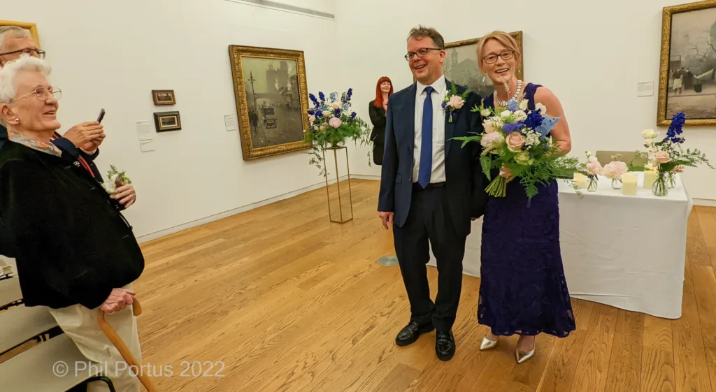 Three people smile for the camera amidst artwork in a room with wooden floors and decorative floral arrangements.