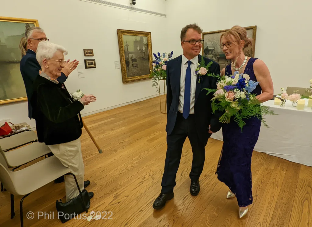 A couple walks hand-in-hand toward a table with floral arrangements in a room lined with framed artwork.
