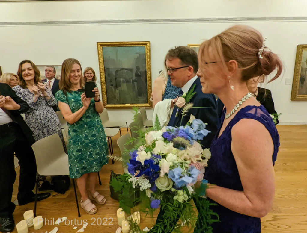 A person in a blue dress carries flowers while walking down an aisle lined with onlookers and framed artwork.