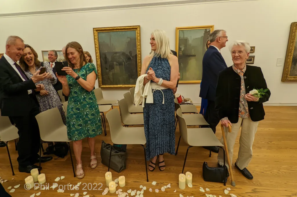 A group of people applaud while standing in a room with paintings on the walls and scattered flower petals on the floor.