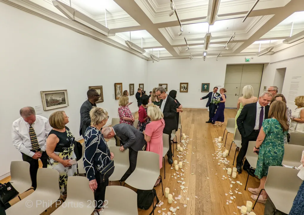 A couple exchanges vows surrounded by guests in a room with wooden floors and artwork on the walls.