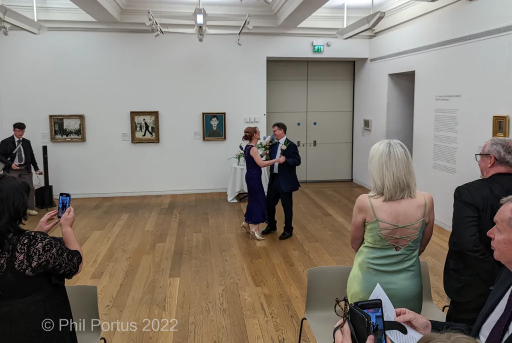 A couple embraces during a ceremony in a room with artwork on the walls and guests seated nearby.