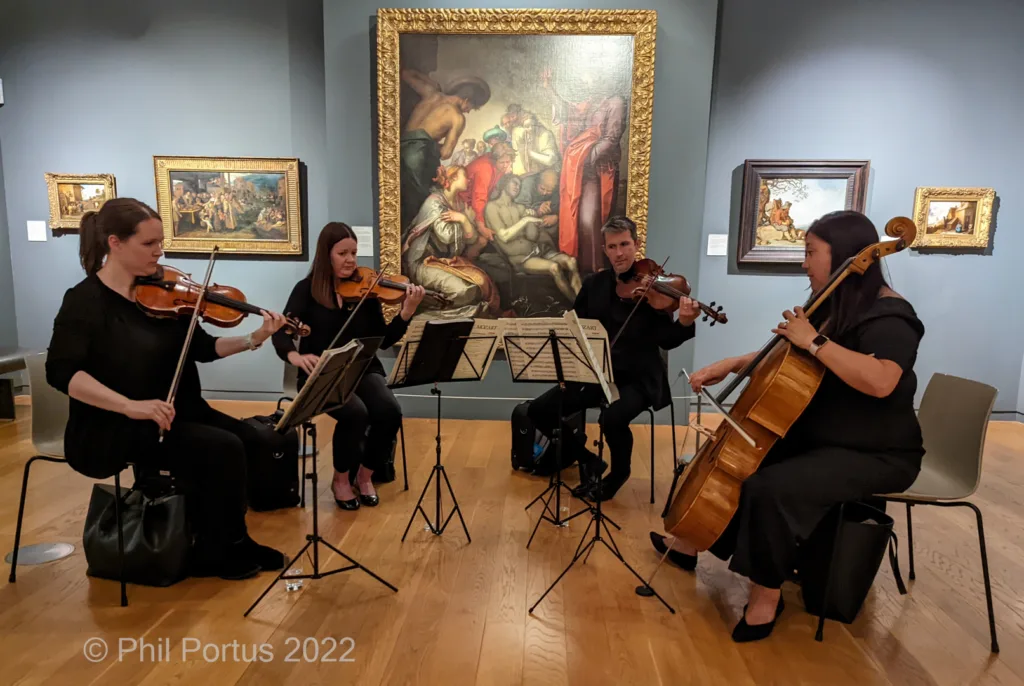 Four musicians seated on chairs perform with string instruments in a room with paintings displayed on the walls.