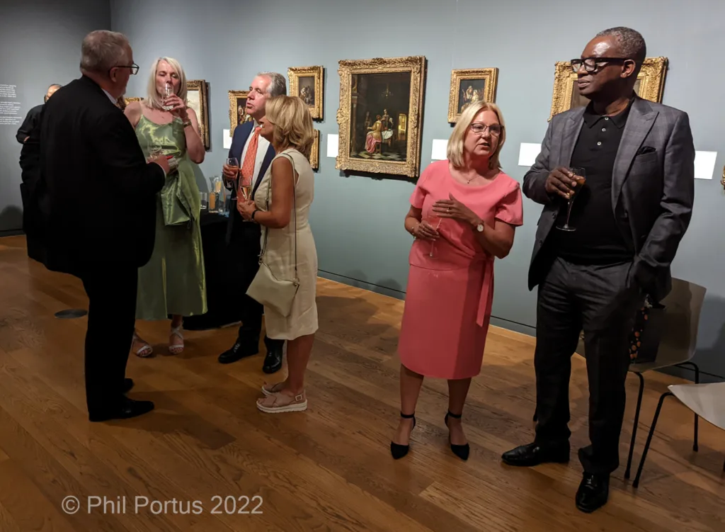 Several people in formal attire converse while holding drinks at an event with framed artwork on the walls.