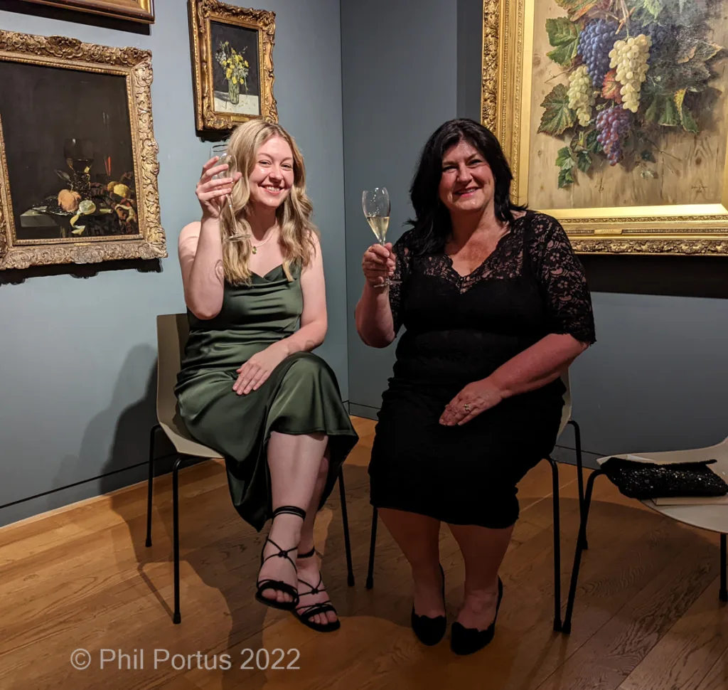 Two women seated on chairs raise glasses in a room with several framed paintings on the walls.