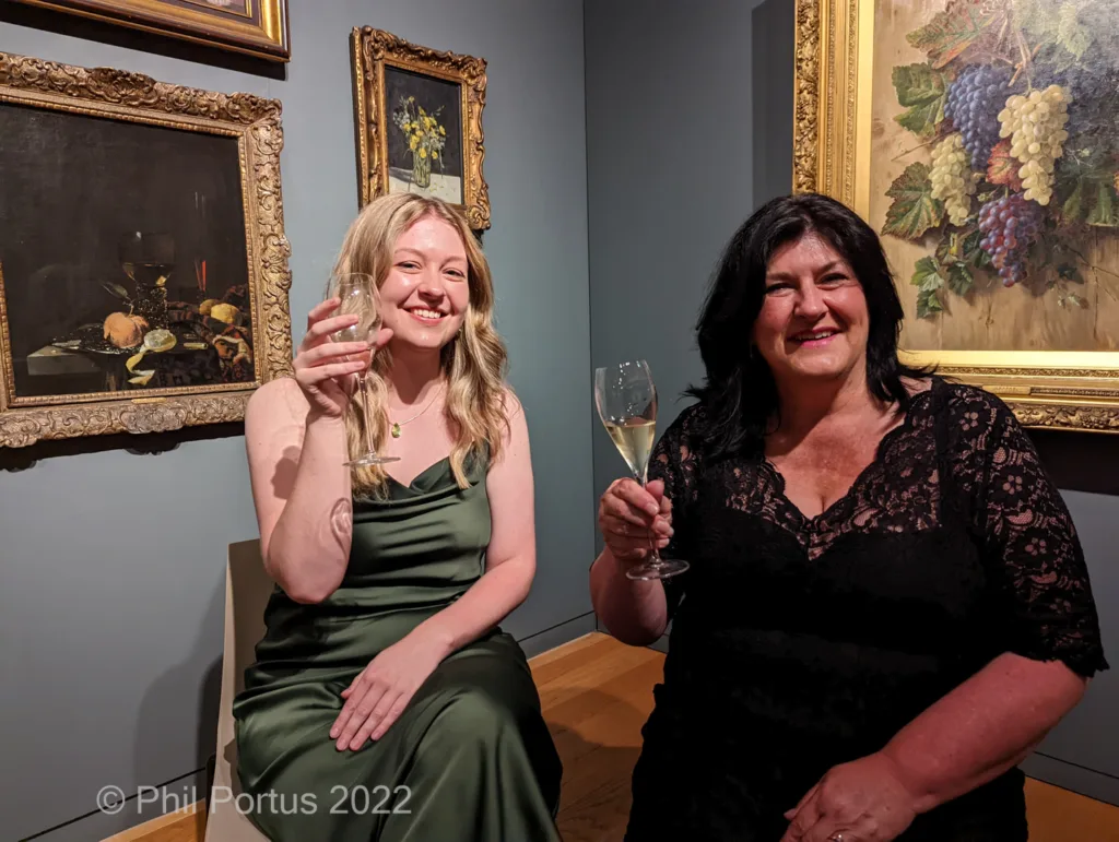 Two women smile while raising glasses of sparkling wine in front of framed still life paintings.