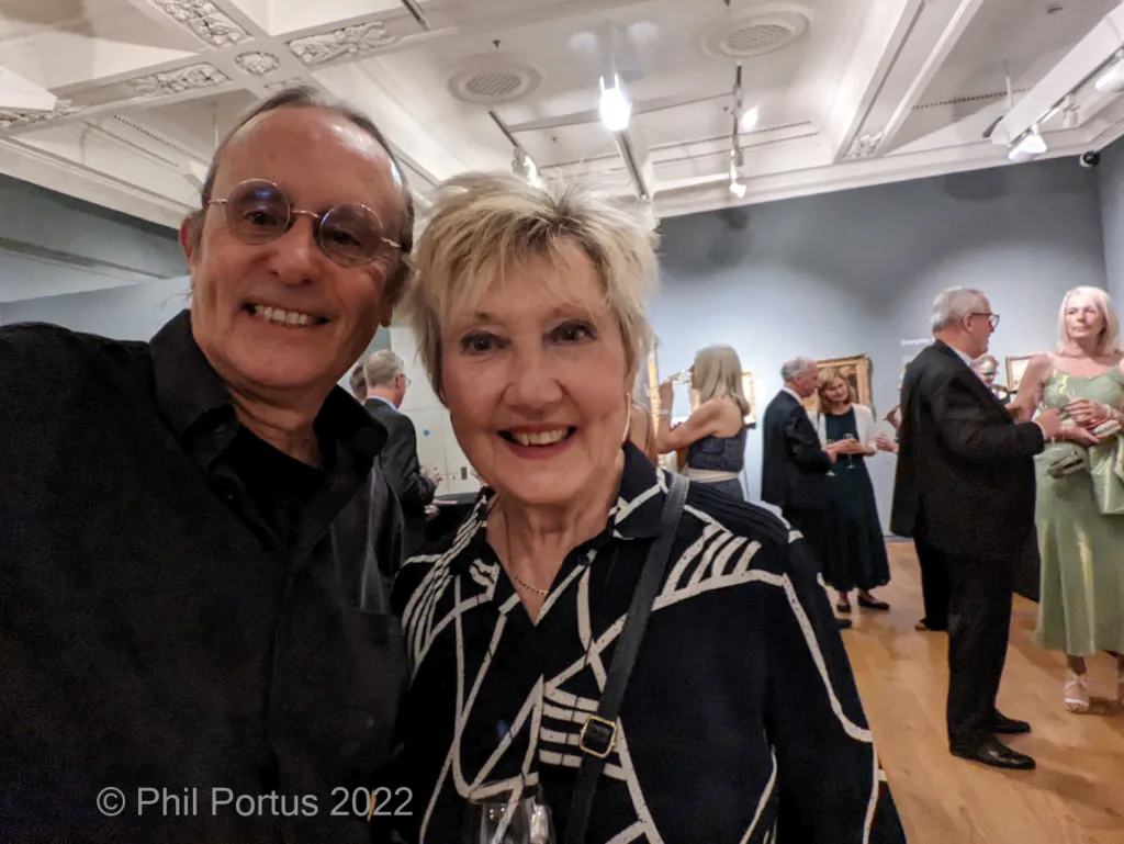 Two people smile for a selfie indoors in a room with high ceilings and artwork on the walls.
