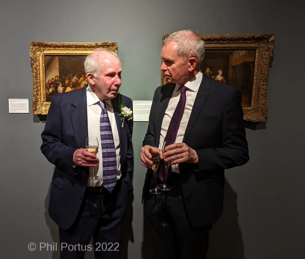 Two older men in suits converse while holding drinks at an event with framed paintings on the wall.