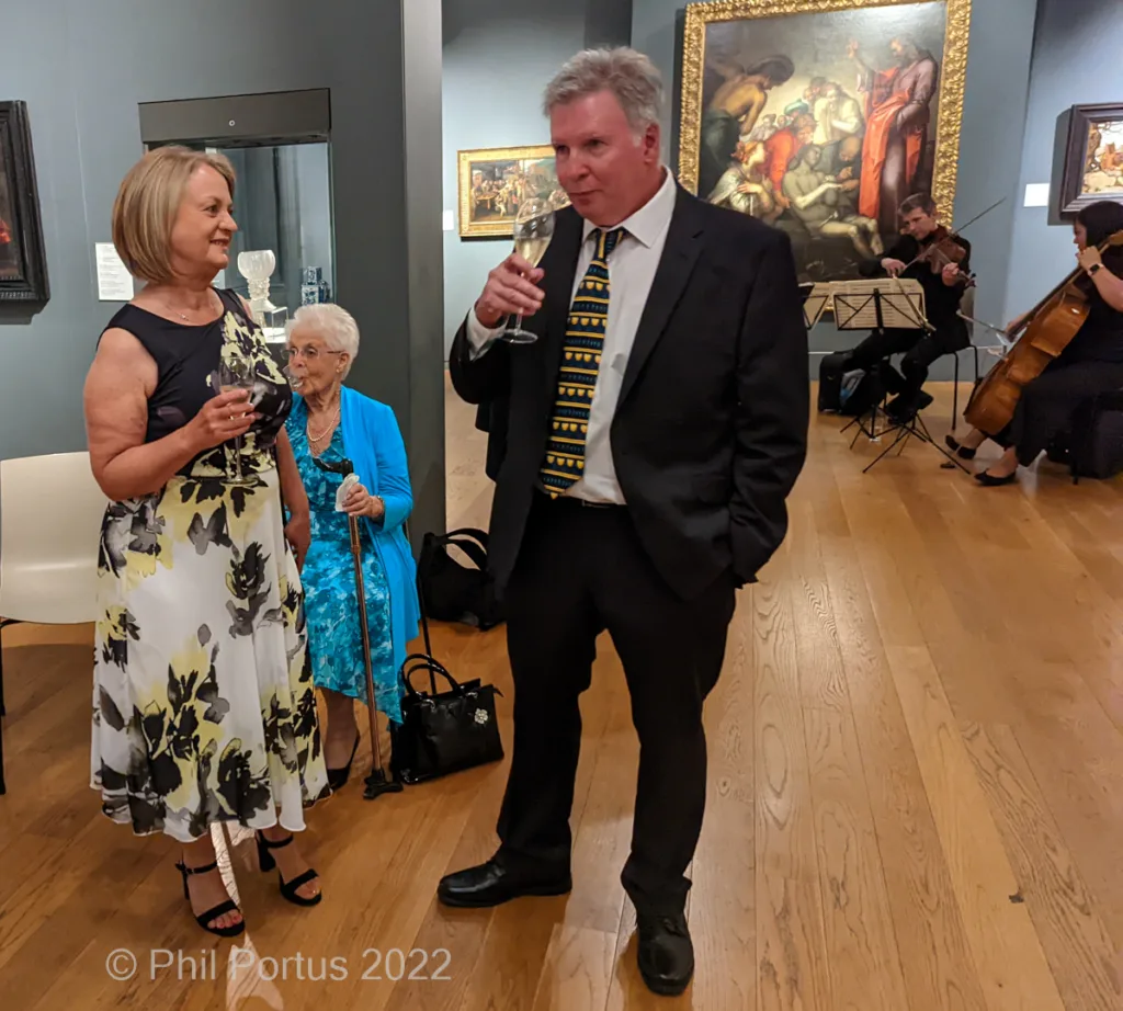 Three people stand on a wooden floor in what appears to be an art gallery, holding glasses.