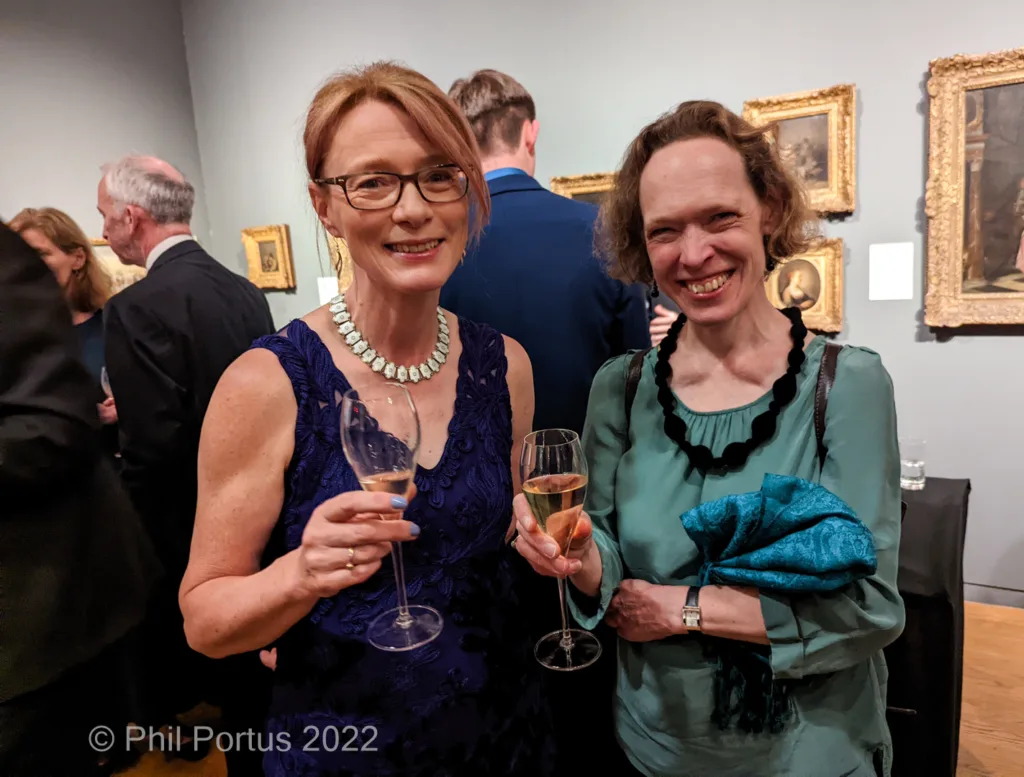 Two women smile while holding glasses of champagne at an event with artwork on the walls.