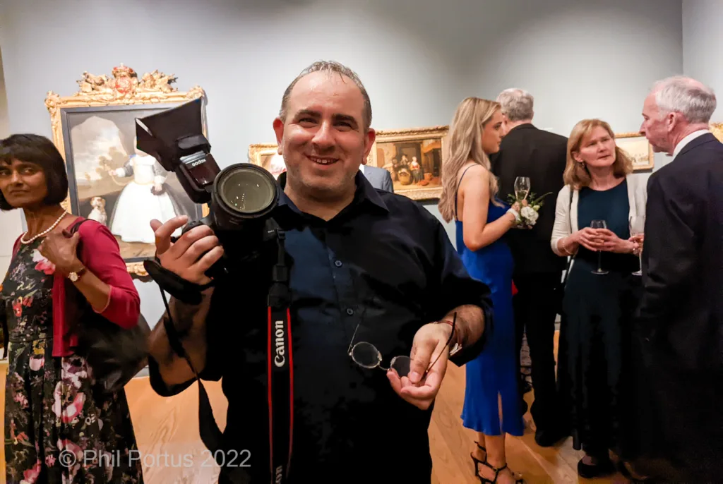 A man holding a camera and light equipment smiles while surrounded by formally dressed people in an ornate room.