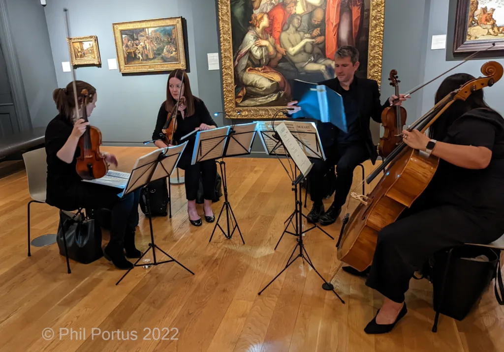 Four musicians perform classical music surrounded by framed paintings in a room with wooden floors.
