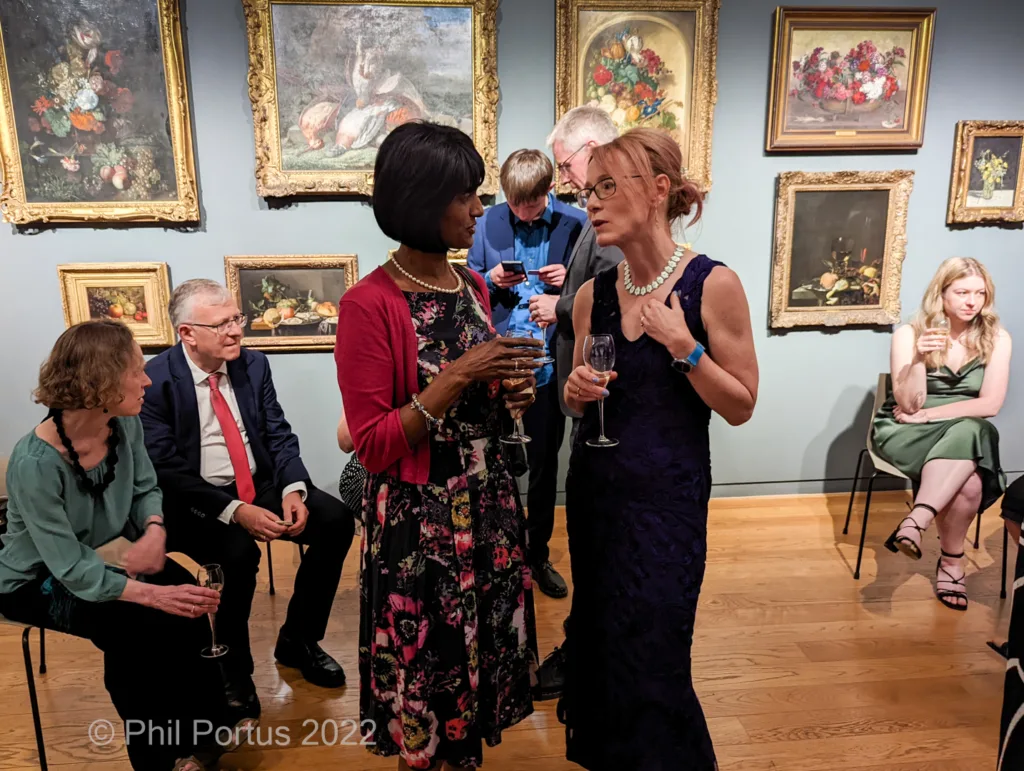 Two women converse while holding glasses at an event with numerous framed paintings on the walls.