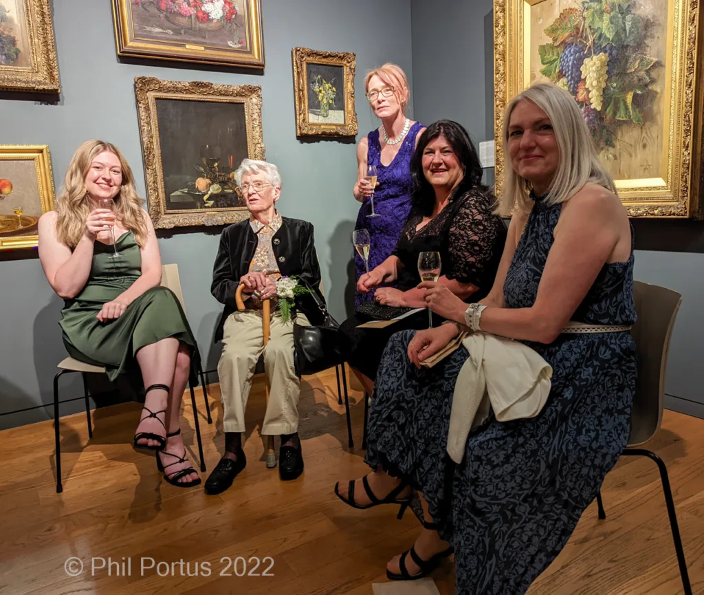 Six people sit on chairs amidst framed paintings in a room with dark walls and wooden floors.