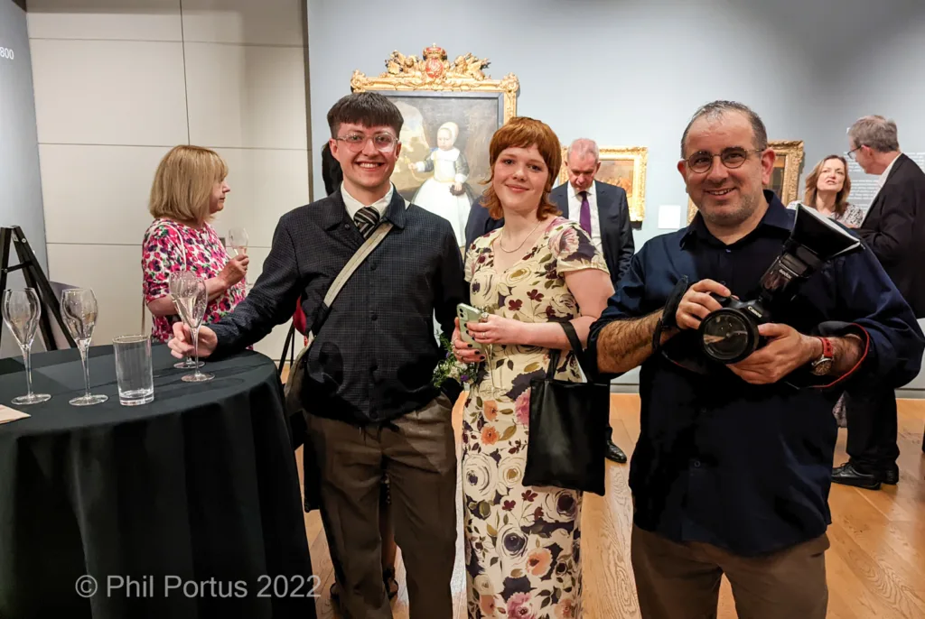 Four people pose near a table with glasses at an event featuring artwork on the wall.