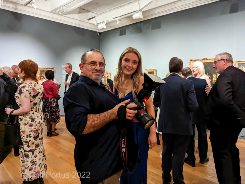 Two people pose with a camera in a brightly lit room filled with other attendees and framed artwork.