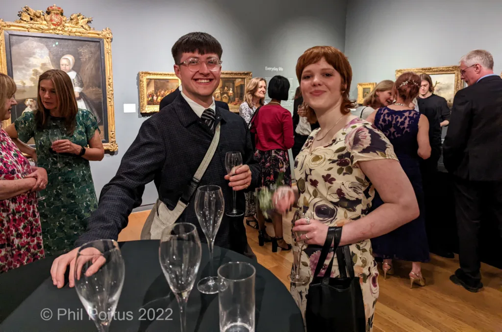Two people holding champagne flutes stand near a table at an event with several paintings on the walls.