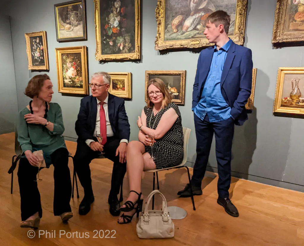 Four people sit and stand amidst framed paintings on a wall in a room with wooden flooring.