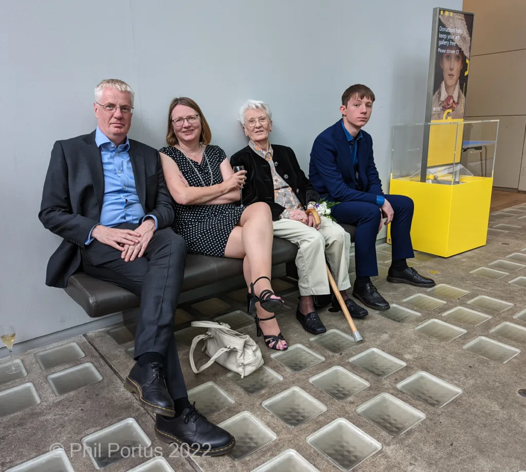 Four people sit on a bench inside a modern building with glass flooring and a yellow display case.