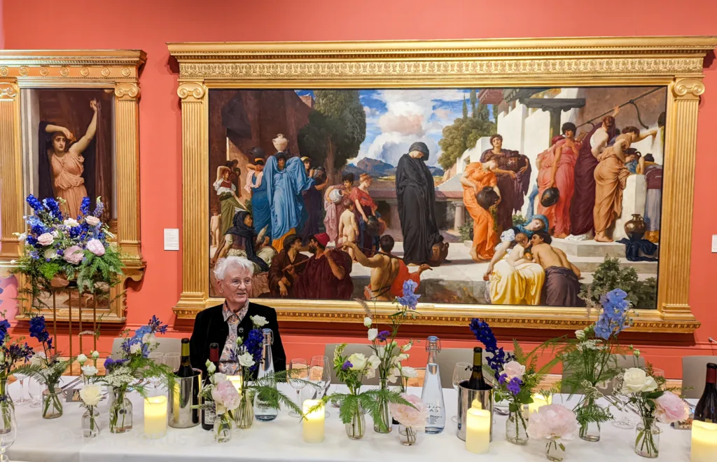A man stands before a long table decorated with flowers and candles, set against large framed paintings.