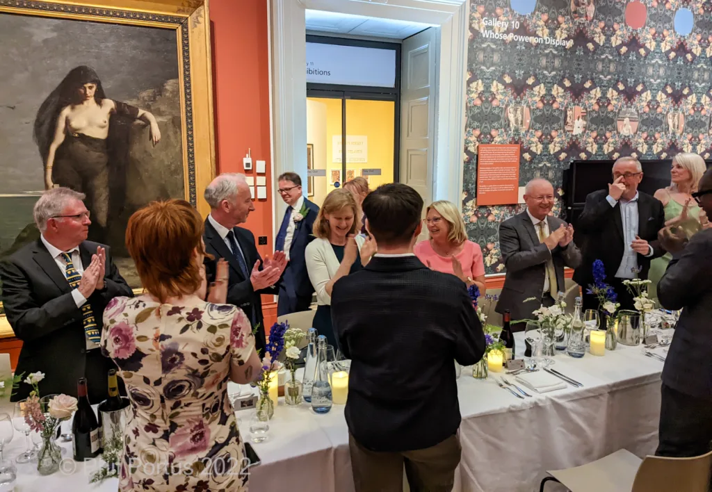 A group of formally dressed people applaud near a long table set for an event in a gallery.