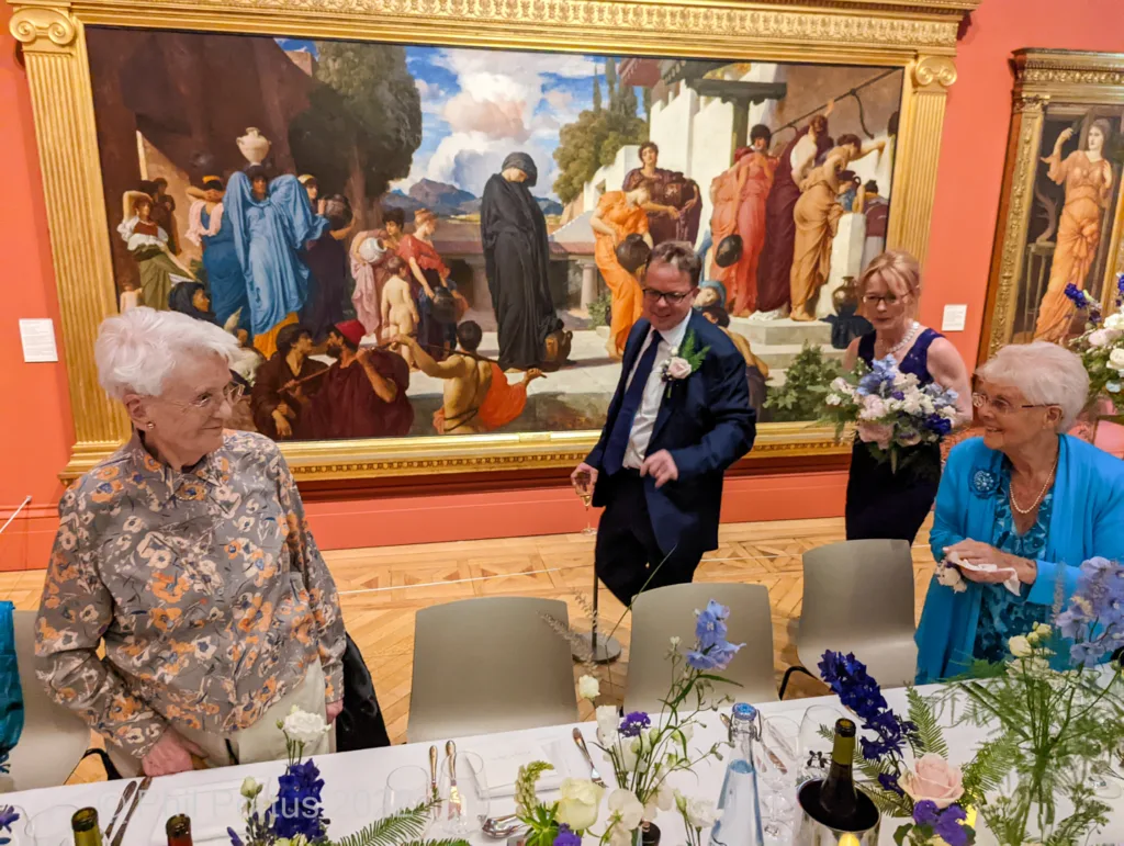 Two older women sit at a table decorated with flowers near a large framed painting and a man in a suit walks past.