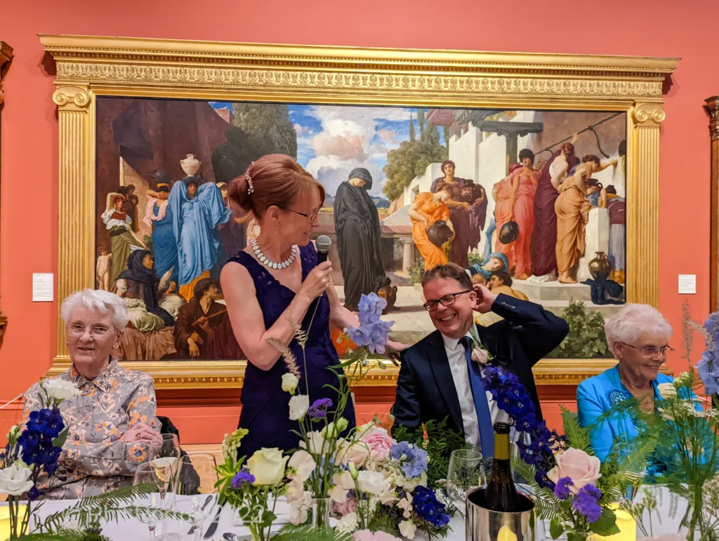 Three people sit at a table adorned with flowers in front of a large framed painting.