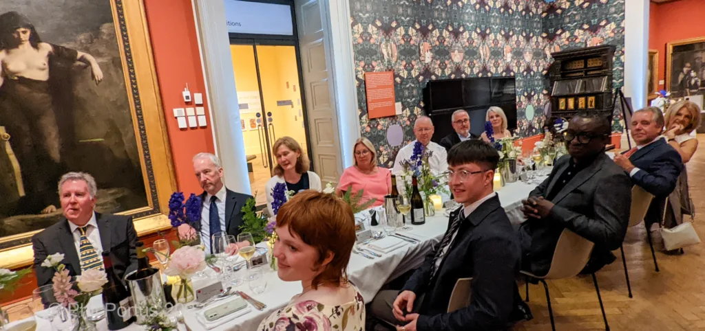 A group of formally dressed people sit at tables set for a dinner event in a room with artwork.