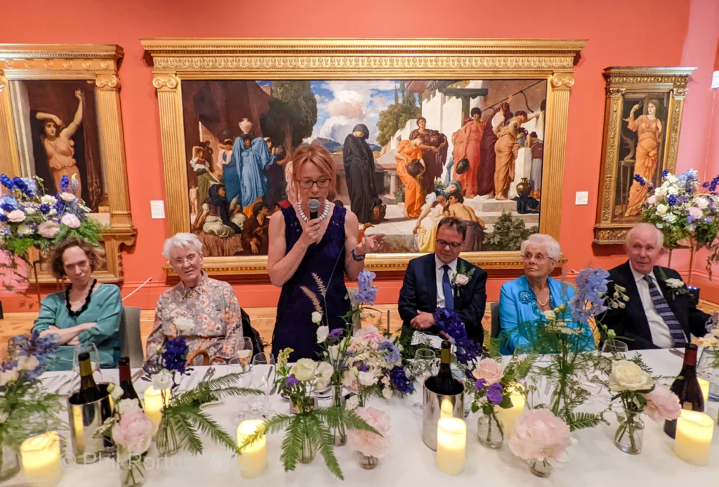 A group gathers around a long table decorated with flowers and candles in a room featuring large framed paintings.